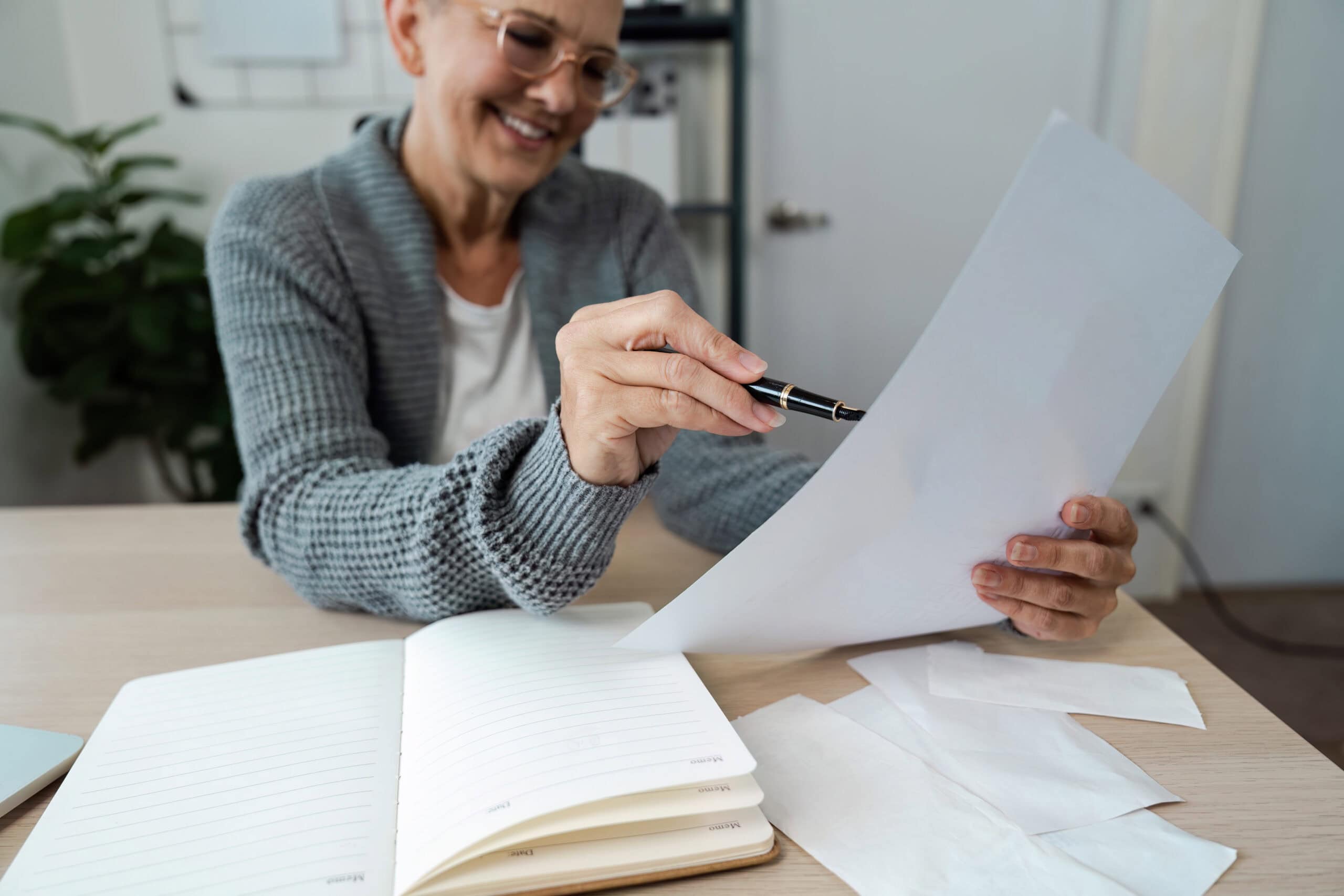 Elderly woman smiling while examining financial paper at her home office desk, surrounded by a notebook and receipts.